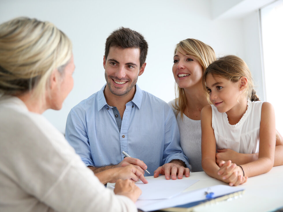 Eine Beraterin sitzt gegenüber einer Familie bestehend aus Mutter, Vater und Kind, welche alle glücklich aussehen.