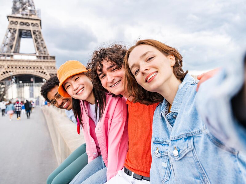 Vier junge Personen lächeln für ein Selfie in die Kamera. Im Hintergrund ist der Eiffelturm zu erkennen.