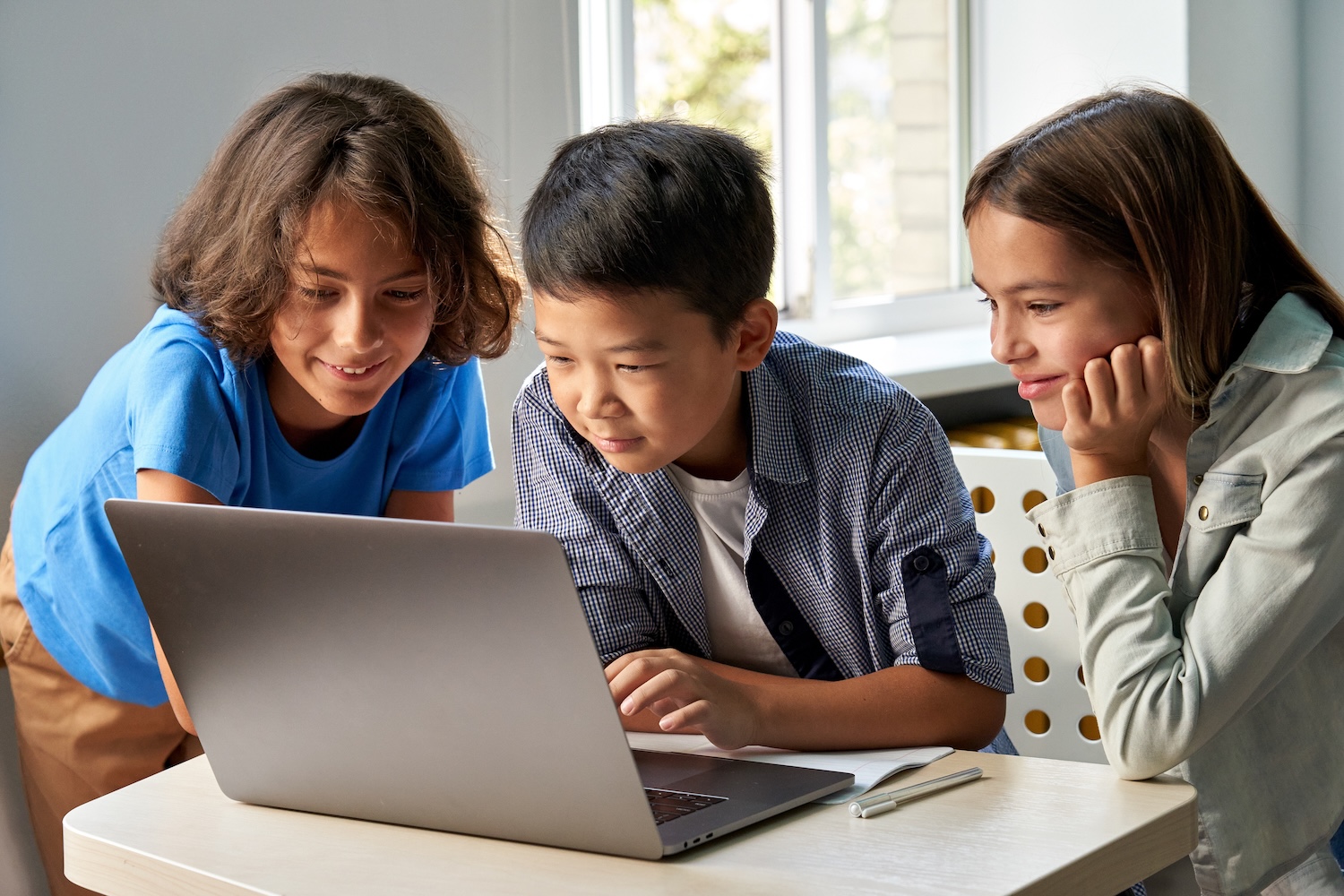 Drei Kinder sitzen gemeinsam an einem Tisch und schauen interessiert auf den Bildschirm eines Laptops.