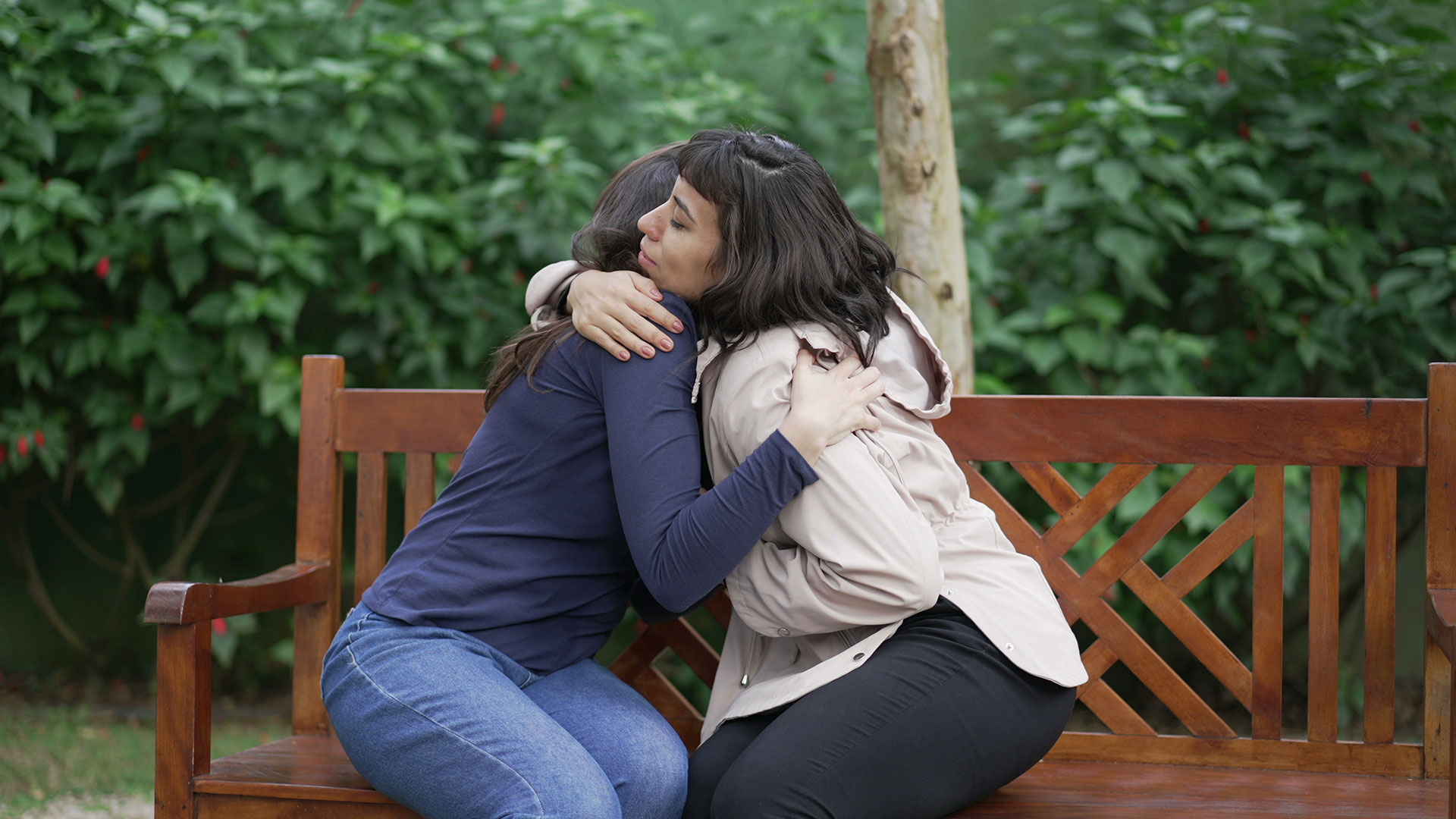 Zwei Frauen umarmen sich tröstend auf einer Bank in der Natur, im Hintergrund eine Wiese und eine Hecke.