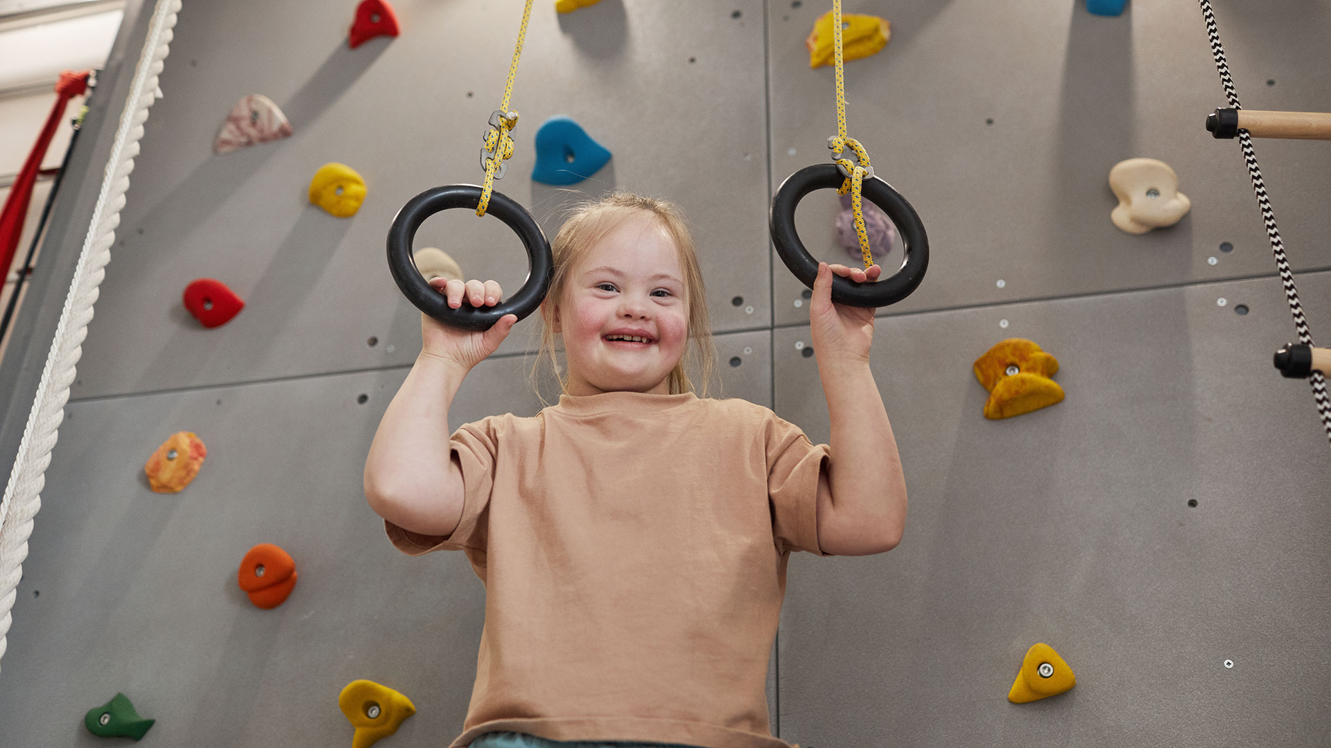 Ein lächelndes Mädchen mit Down Syndrom greift Ringe vor einer Kletterwand