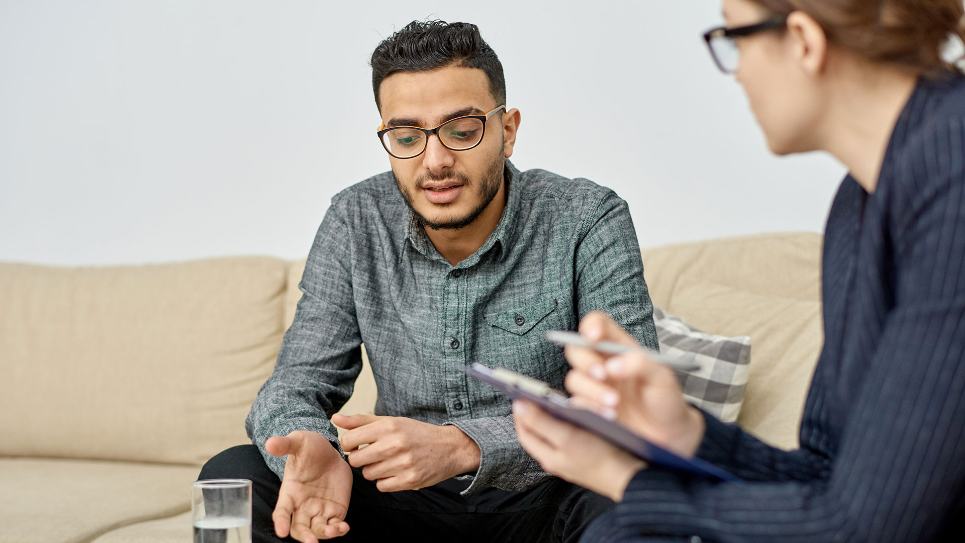 Junger Mann mit Brille sitzt auf einer Couch, neben ihm auf einem Stuhl sitzt eine Frau, die sich Notizen macht