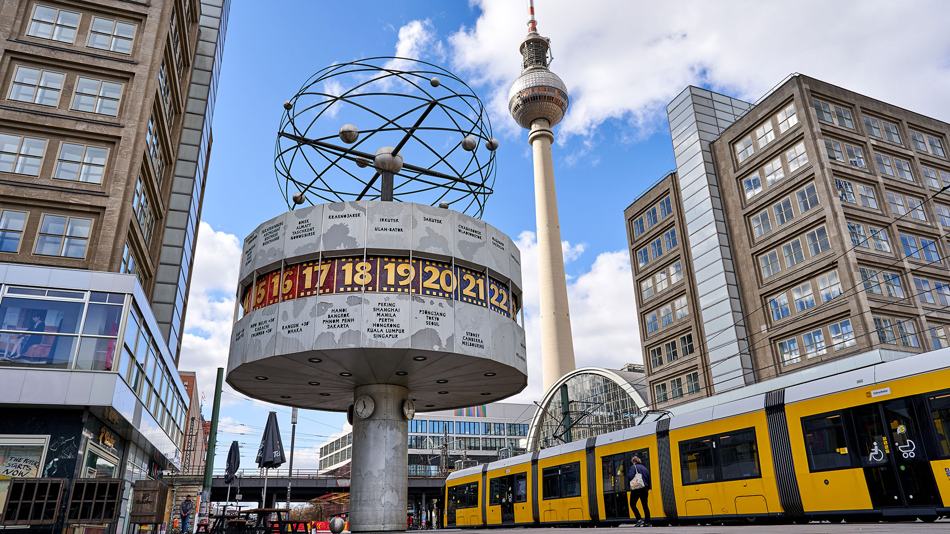 Die Weltzeituhr auf dem Berliner Alexanderplatz mit Blick auf den Fernsehturm. Durch das Bild fährt eine Straßenbahn der BVG.