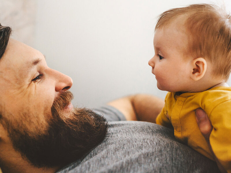 Ein Vater schaut lächelnd sein auf dem Bauch liegendes Baby an.