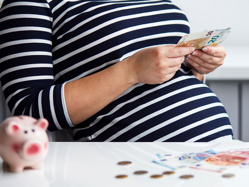 Eine Frau mit Babybauch sitzt am Tisch und zählt Euroscheine, auf dem Tisch steht zudem eine rosa Spardose in Form eines Schweines 