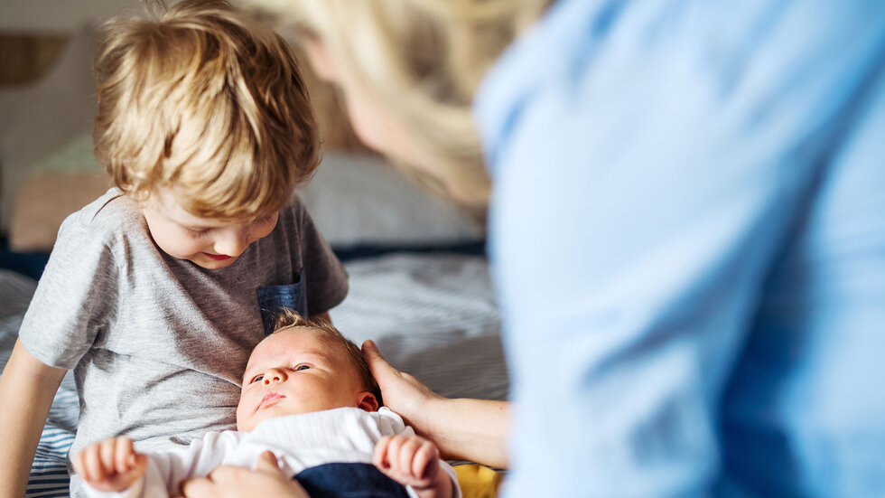 Ein Kleinkind hat ein Baby auf dem Schoß liegen, dass von einer Frau gestreichelt wird.