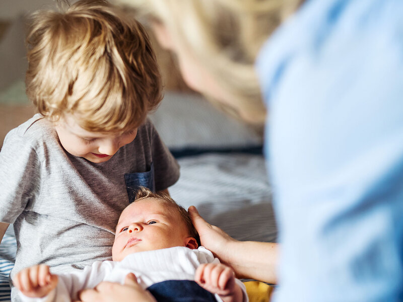 Ein Kleinkind hat ein Baby auf dem Schoß liegen, dass von einer Frau gestreichelt wird.